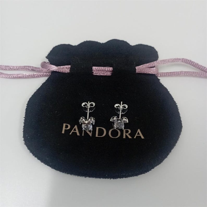 May include: A pair of silver stud earrings with a clear, square-cut gemstone. The earrings are displayed on a black velvet pouch with the brand name "PANDORA" printed in white. The pouch has a pink drawstring.
