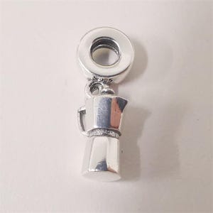 May include: A silver charm depicting a small coffee maker. The charm has a loop at the top for attachment and is made of polished metal. The coffee pot charm is a jewelry accessory, suitable for a bracelet or necklace.