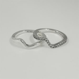 May include: Two silver rings with a wave design. One ring is plain silver, while the other features a row of small, clear stones along the wave. The rings are interlocking, creating a layered effect. The word "PANDORA" is visible on one ring.