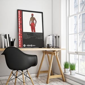 May include: A framed poster featuring Tyler, the Creator, with the text "TYLER THE CREATOR DON'T TAP THE GLASS" in red. The poster displays a photo of Tyler in red trousers and a gold chain. The poster is on a wooden desk.