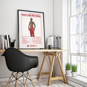 May include: Framed poster featuring Tyler, The Creator, with the text "Don't Tap The Glass" in red. The image shows Tyler in red trousers and a red hat. The poster is on a wooden desk with books, a black chair, and a window.