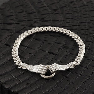 May include: Silver-toned bracelet featuring a curb chain design. The bracelet's clasp showcases two detailed hand sculptures holding a circular ring. The jewelry is displayed against a dark, textured surface, highlighting the metallic sheen.