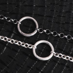 Könnte beinhalten: Nahaufnahme von zwei silberfarbenen Halsketten. Jede Halskette hat einen großen, kreisförmigen Anhänger, der mit einer filigranen Kette verbunden ist. Die Ketten bestehen aus kleinen, miteinander verbundenen Gliedern. Der Hintergrund ist eine strukturierte, dunkle Oberfläche.