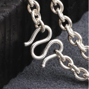 May include: Close-up of a silver chain with a curved clasp. The chain links are round and connected, with a shiny, reflective surface. The clasp is in the shape of an S, and the background is dark.