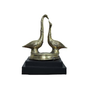 Può includere: Una scultura in ottone di due oche che si fronteggiano, con colli lunghi e ali dettagliate. Le oche sono montate su una base circolare, che poggia su una base rettangolare nera. La scultura ha una finitura lucida.