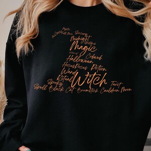 Könnte beinhalten: Schwarzes Sweatshirt mit einem orangefarbenen Hexenhut-Design. Der Hut ist mit Wörtern wie "Magic", "Witch" und "Halloween" gefüllt. Das Sweatshirt hat einen Rundhalsausschnitt und lange Ärmel.