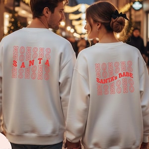May include: Two white sweatshirts with the text "HOHOHO SANTA HOHOHO" and "HOHOHO SANTA'S BABE HOHOHO" in red. The sweatshirts are being worn by a couple walking down a street decorated with Christmas lights.