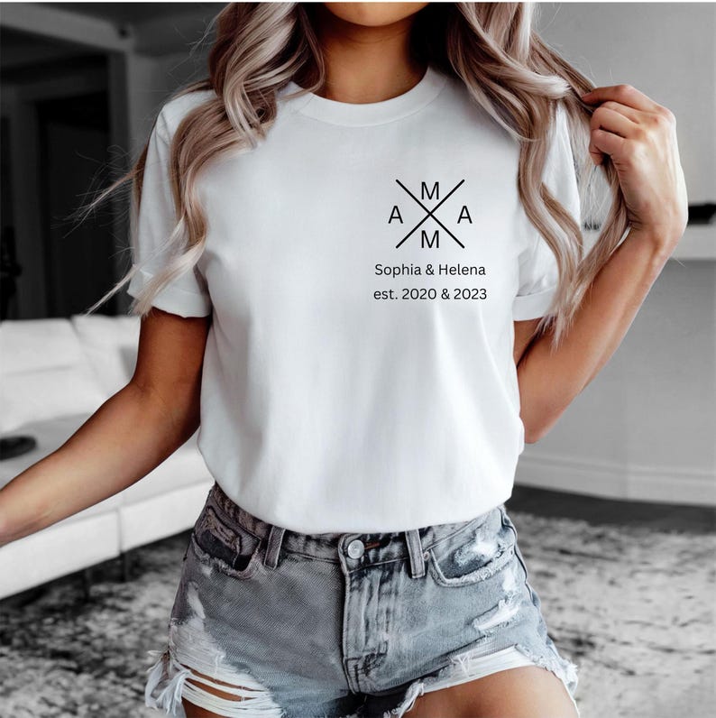 May include: White t-shirt with a black graphic design featuring the letters "A" and "M" crossed, with the text "Sophia & Helena est. 2020 & 2023" below. The shirt has short sleeves and a crew neck.