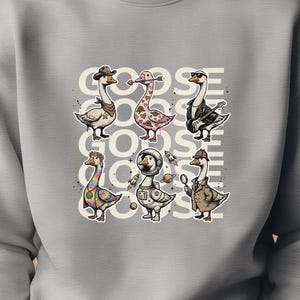 May include: A light gray sweatshirt featuring a graphic of several cartoon geese, each with unique designs and accessories. The word "GOOSE GOODS" is printed above the geese. The sweatshirt is made of a soft material.