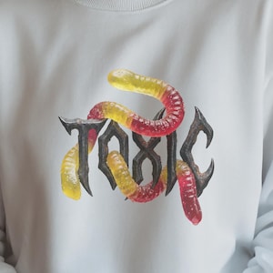 May include: White sweatshirt with the word "TOXIC" in a gothic font, with colorful gummy worms. The worms are yellow, red, and orange. The sweatshirt has long sleeves and a crew neck.