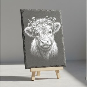 May include: A rectangular slate sign featuring a detailed white illustration of a Highland cow wearing a floral crown. The sign is displayed on a small wooden easel, showcasing the artwork's intricate details and rustic charm.