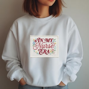 Pode incluir: Camisola branca com um patch retangular bordado com o texto "IN MY Nurse ERA" em itálico. O patch é decorado com bordados florais em tons de rosa, azul e amarelo. A camisola tem gola redonda e mangas compridas.
