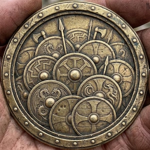 May include: A bronze-colored, circular medallion featuring a detailed relief of Viking shields, axes, and spears. The design includes various shield patterns, some with dragon motifs. The outer rim has a series of rivets, giving it a rugged, historical appearance.