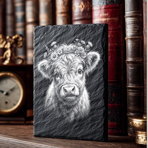 May include: A black rectangular slate featuring a detailed white illustration of a Highland cow's face, adorned with a floral crown. The cow's fur is intricately rendered, and the slate has a textured surface. The image is set against a backdrop of antique books.