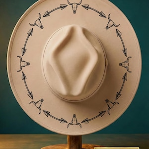 Puede incluir: Un sombrero de vaquero beige con ala ancha, adornado con un patrón repetido de calaveras de toro negras y flechas. El sombrero se exhibe sobre un soporte de madera. Un pequeño letrero dorado en la parte inferior dice "Descarga digital: PNG + PDF".