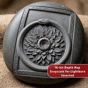 May include: A dark gray, round stone with a carved design. The design features a snake eating its tail around a flower, framed within a square. The text "16-bit Depth Map Grayscale for Lightburn Inverted" is visible on the bottom right.