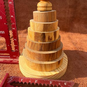 Op de afbeelding: Een houten stapelspeelgoed met een lichtbruine afwerking. Het speelgoed bestaat uit verschillende cirkelvormige stukken van verschillende grootte, gestapeld om een toren te vormen, bekroond met een decoratief stuk. Een rode meetvierkant staat op de voorgrond.