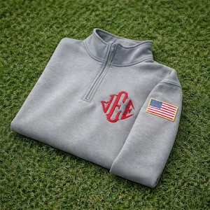 Op de afbeelding: Een grijze quarter-zip sweatshirt met een rood monogram en een Amerikaanse vlag patch op de mouw. De sweatshirt is gemaakt van een zacht materiaal en ligt op groen gras.