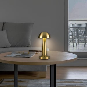 May include: A gold-colored table lamp with a mushroom-shaped shade. The lamp has a cylindrical base and stem, and it is placed on a round wooden table. The lamp is illuminated, casting a warm glow. The background includes a gray sofa and a window.
