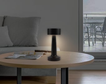 May include: A black, modern table lamp with a cylindrical shade and a slender, conical base. The lamp is illuminated, casting a warm glow. It sits on a round wooden table, suggesting a contemporary interior design.
