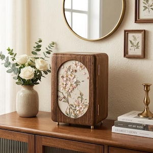 Puede incluir: Un joyero rectangular de madera con acabado en nogal oscuro y un panel ovalado central que presenta bordados florales en relieve en tonos rosa, blanco y verde. El joyero se apoya sobre cuatro pequeñas patas de color dorado y tiene bisagras doradas.