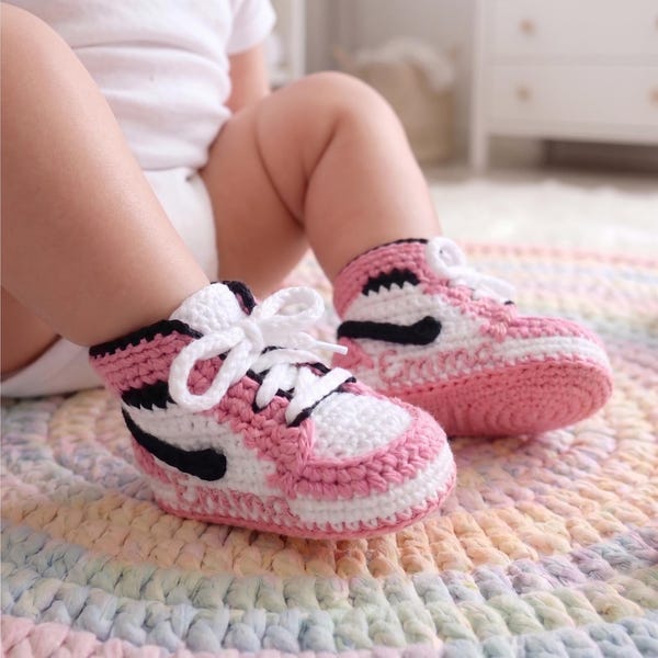 May include: A pair of handmade crochet baby booties designed to resemble high-top sneakers. The booties feature a color-blocked pattern in white, soft pink, and black yarn, complete with white laces and the name Emma stitched on the side panels.