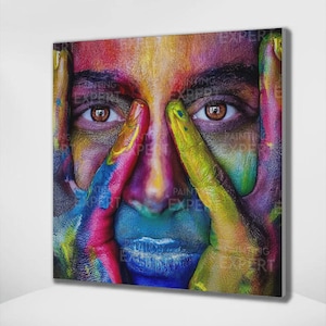 Puede incluir: Una pintura en primer plano de un rostro con pintura colorida que cubre la piel. Los ojos son marrones y los labios azules. Las manos están cerca de la cara, con los dedos pintados en varios colores, incluyendo amarillo, rosa y azul.