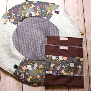 Puede incluir: Una alfombra de juego redonda multicolor con un círculo central gris y un bolsillo de tela marrón con dos compartimentos. La alfombra tiene un patrón floral con mariposas y pájaros. El bolsillo tiene una etiqueta que dice "LA GRANDE AVENTURE".