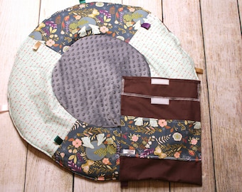 May include: A round, multi-colored play mat with a gray center circle and a brown fabric pocket with two compartments. The mat has a floral pattern with butterflies and birds. The pocket has a label that says "LA GRANDE AVENTURE".