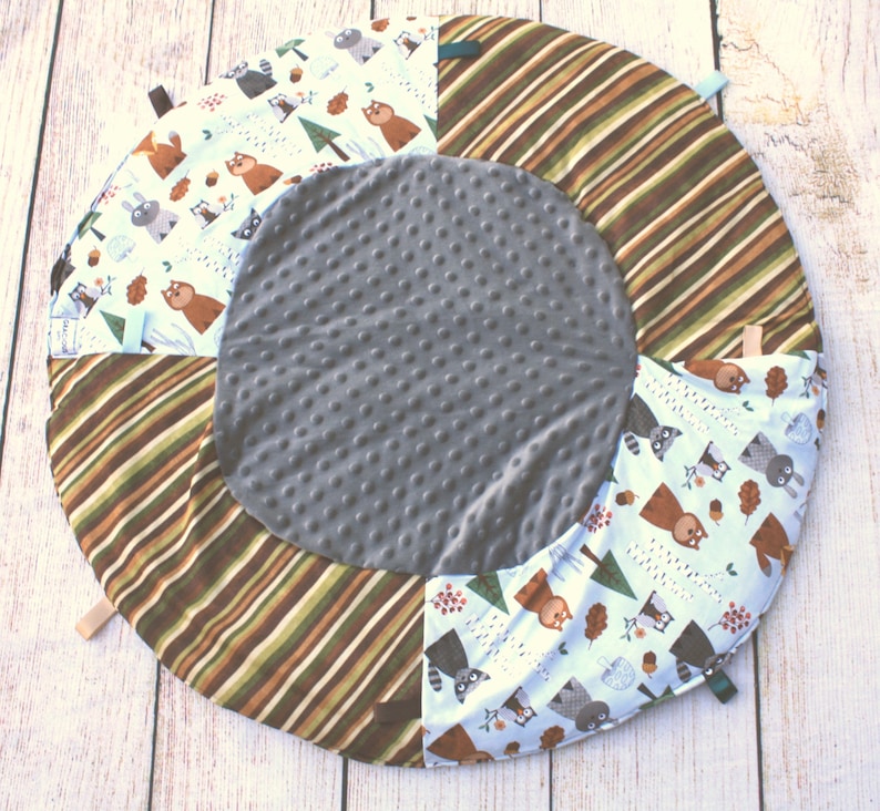 Puede incluir: Una alfombra de juego redonda, gris y marr&oacute;n con estampado de animales blanco y marr&oacute;n. La alfombra tiene un centro suave y texturizado y un borde exterior a rayas.