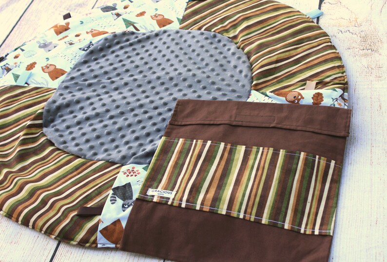 Puede incluir: Una alfombra de juego redonda multicolor con un centro acolchado gris. La alfombra tiene un bolsillo de tela marr&oacute;n con un panel de tela a rayas. La alfombra presenta un tema de bosque con im&aacute;genes de osos, mapaches y &aacute;rboles.