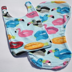 Könnte beinhalten: Zwei Baby-Lätzchen mit hellblauem Hintergrund mit Tukanen, Flamingos und Fruchtscheiben. Die Lätzchen haben eine weiße Bordüre und ein Etikett mit der Aufschrift "Gracious Gifts".