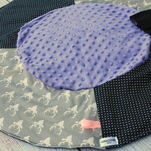 May include: A multi-colored baby play mat with a purple, textured center circle. The mat has a black and white polka dot pattern, a gray fabric with white deer silhouettes, and a black fabric with a white tag that reads "Gracious Gifts".