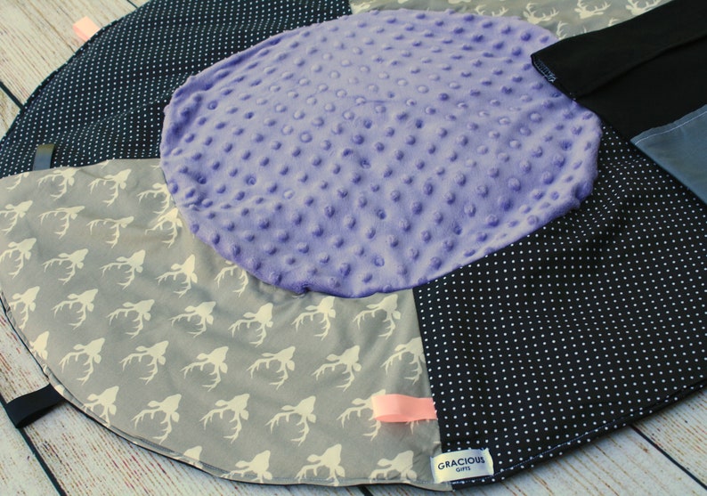May include: A multi-colored baby play mat with a purple, textured center circle. The mat has a black and white polka dot pattern, a gray fabric with white deer silhouettes, and a black fabric with a white tag that reads "Gracious Gifts".