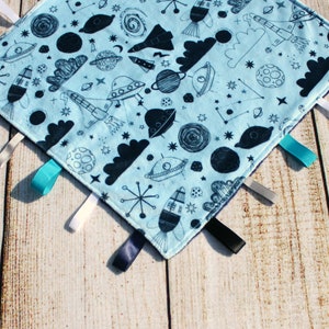 Puede incluir: Una manta de bebé azul con temática espacial. La manta tiene una variedad de ilustraciones en blanco y negro de cohetes, planetas, estrellas y OVNIs. La manta tiene cinco cintas de satén atadas a las esquinas.