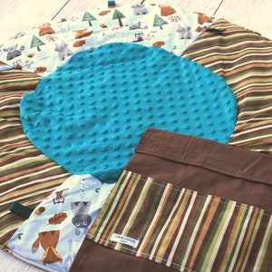 Puede incluir: Una alfombra de juego redonda multicolor con un borde exterior a rayas marr&oacute;n, verde y blanco. El centro de la alfombra presenta un c&iacute;rculo turquesa texturizado. Un bolsillo de tela marr&oacute;n con un bolsillo a rayas y el texto "Gracious" est&aacute; unido a la alfombra.