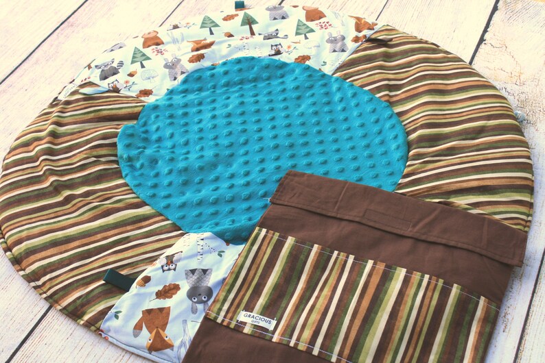 Puede incluir: Una alfombra de juego redonda multicolor con un borde exterior a rayas marr&oacute;n, verde y blanco. El centro de la alfombra presenta un c&iacute;rculo turquesa texturizado. Un bolsillo de tela marr&oacute;n con un bolsillo a rayas y el texto "Gracious" est&aacute; unido a la alfombra.