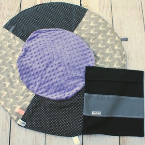 May include: A round, multi-colored baby play mat with a purple center, black and white polka dot fabric, and a gray fabric with white deer silhouettes. A black fabric pouch with a gray stripe and the word "Remarkable" is included.