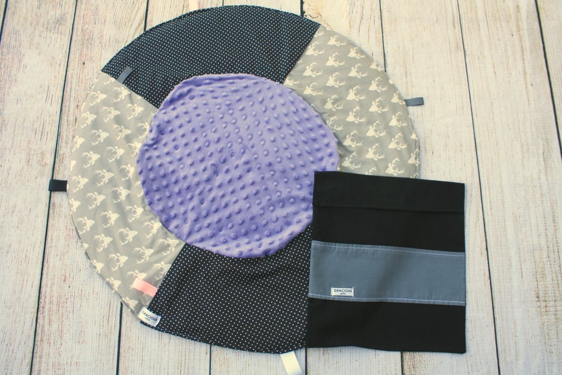 May include: A round, multi-colored baby play mat with a purple center, black and white polka dot fabric, and a gray fabric with white deer silhouettes. A black fabric pouch with a gray stripe and the word "Remarkable" is included.