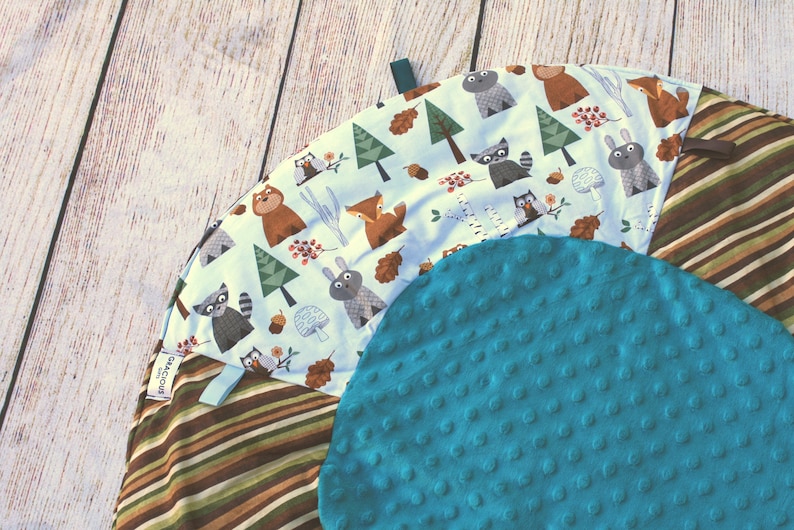 Puede incluir: Una alfombra de juego redonda y multicolor para beb&eacute;s con un centro texturizado azul. El anillo exterior presenta un patr&oacute;n de rayas marrones y blancas. La secci&oacute;n central presenta un fondo blanco con un patr&oacute;n repetitivo de animales del bosque y &aacute;rboles. El texto "Gracious" es visible en la alfombra.