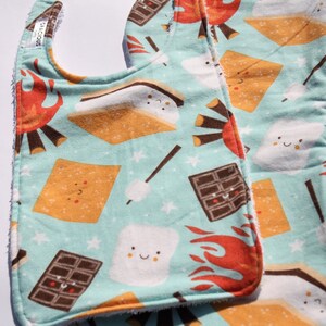 以下が含まれることがあります： 白いテリークロス裏地が付いたターコイズブルーのベビーよだれかけ2枚。よだれかけには、スモア、チョコレートバー、マシュマロ、キャンプファイヤーが繰り返しパターンで描かれています。1枚のよだれかけには「S'mores」という文字が印刷されています。