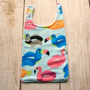 Könnte beinhalten: Ein blaues und weißes Baby-Lätzchen mit einem tropischen Druck mit Flamingos und Tukanen. Das Lätzchen hat einen Halsausschnitt und ein Etikett mit der Aufschrift "Gracious Gifts".