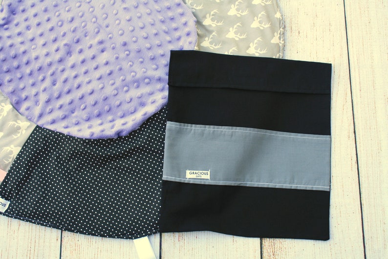 May include: A black fabric storage bag with a gray stripe and the text "Gracious Gifts" on a white label. The bag is lying on a white wooden surface.