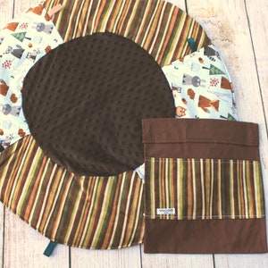 Puede incluir: Una alfombra de juego marr&oacute;n y blanca con tem&aacute;tica de animales del bosque. La alfombra tiene un c&iacute;rculo central marr&oacute;n y un anillo exterior a rayas. Viene con una bolsa de almacenamiento marr&oacute;n con un bolsillo a rayas. La bolsa tiene una etiqueta blanca con el texto "Gracious".