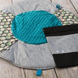Puede incluir: Una alfombra de juego redonda, gris y blanca con un círculo central turquesa y un patrón de calavera blanco y negro. La alfombra tiene una bolsa de almacenamiento negra con la palabra "Gracious" impresa en ella.