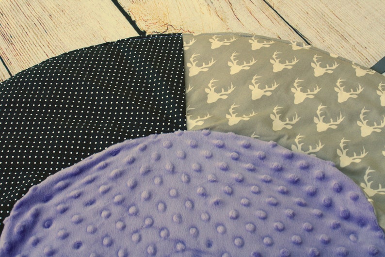 May include: A round, multi-colored play mat with three different fabric patterns. One section is black with white polka dots, another is gray with white deer silhouettes, and the third is a soft purple with a textured surface.