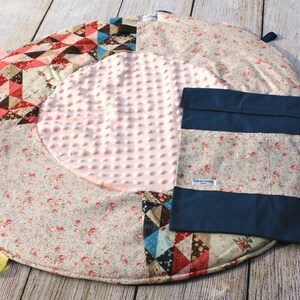 Puede incluir: Una alfombra de juego redonda multicolor con un círculo central rosa texturizado. La alfombra tiene un diseño de patchwork con patrones florales y geométricos. Una bolsa de tela azul con un patrón floral blanco y el texto "GRACE KIDS" está unida a la alfombra.