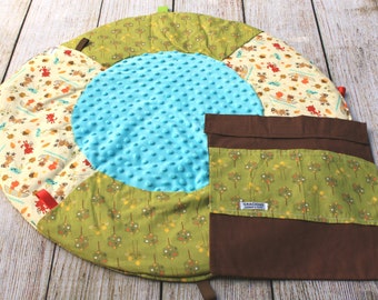 May include: A round, multi-colored play mat with a blue, padded center. The mat has a brown pocket with the text "Gracious" on it. The mat is made of fabric with a variety of patterns, including trees, animals, and leaves.