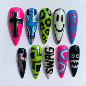 May include: A set of ten long, pointed artificial nails with various designs. The designs include a cross, a Union Jack, leopard print, a smiley face, and the word "SWAG". Colors include pink, blue, green, black, and white.