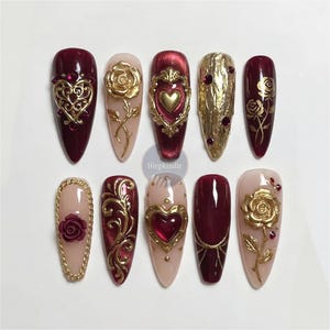 May include: A collection of ten artificial nails in diverse designs. The nails showcase burgundy, gold, and nude hues. Some nails are adorned with gold heart and rose decorations, while others feature elaborate gold filigree patterns.